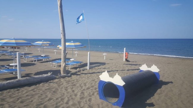 Una spiaggia con ombrelloni, lettini, una bandiera blu alzata e un tunnel-gioco nero sulla sabbia.
