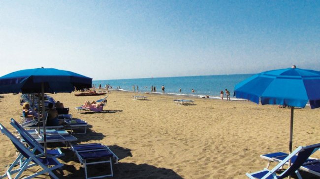 Una spiaggia con ombrelloni blu, sedie a sdraio e persone che camminano sul bagnasciuga.