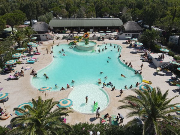 Ampia vista di una piscina affollata con giochi d'acqua. Palme e ombrelloni circondano la zona.