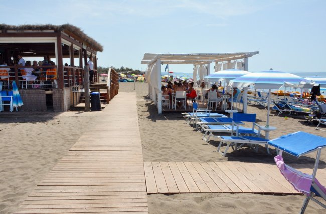 Sdraio e ombrelloni su una spiaggia, vicino a un bar e una sala con tavoli coperti.