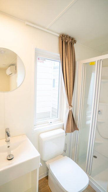 Bagno moderno con lavabo, specchio rotondo, WC e doccia. Finestra con tenda marrone chiusa.
