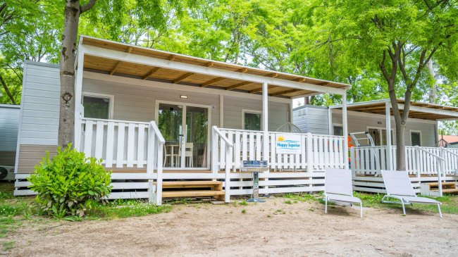 Casa mobile con veranda bianca, sedie a sdraio e alberi intorno.
