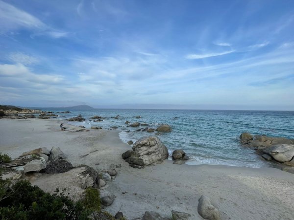 Spiaggia rocciosa con sabbia bianca e mare azzurro sotto un cielo nuvoloso.