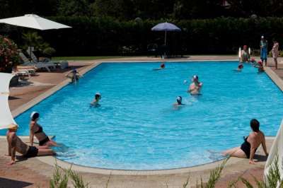 Piscina con bagnanti e ombrelloni, circondata da verde.