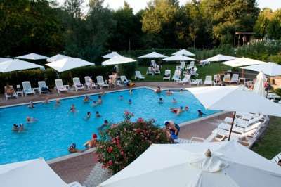 Una piscina affollata con lettini e ombrelloni bianchi. Alberi e vegetazione sullo sfondo.