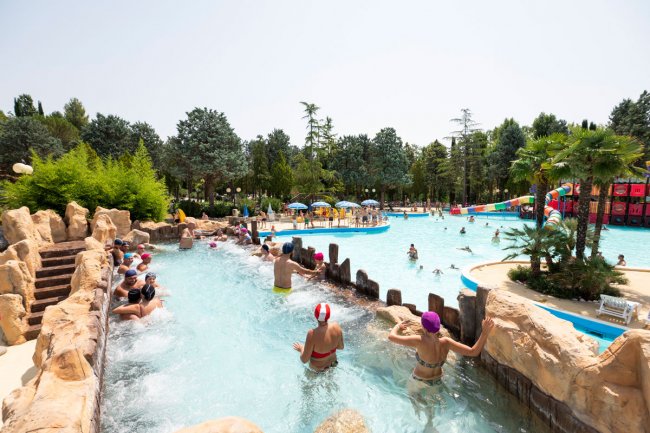 Persone in una piscina con scivolo e alberi circostanti.