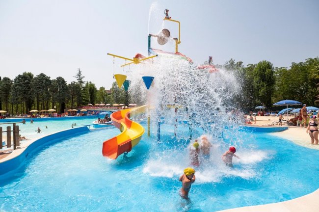 Bambini giocano in piscina con scivolo e spruzzi d'acqua.