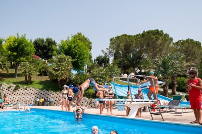 Persona fa un tuffo in piscina. Persone attorno guardano.