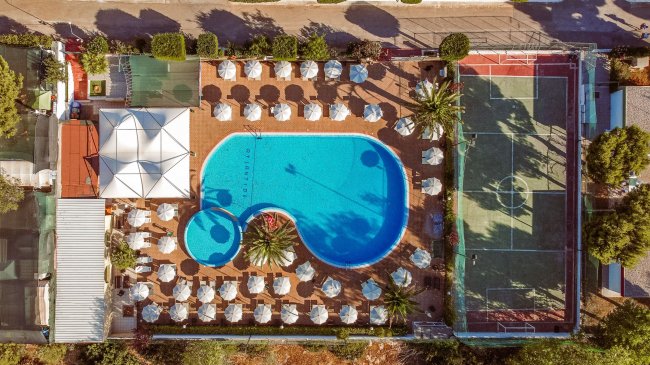 Vista aerea di una piscina circondata da ombrelloni e campo da tennis.