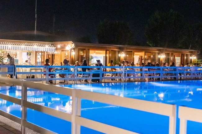 Piscina illuminata di notte con ristorante affollato sullo sfondo.
