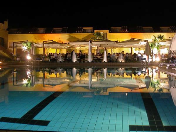 Piscina illuminata di notte con persone cenanti sotto pergole.