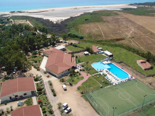 Vista aerea di un complesso turistico con piscina, campi sportivi e mare sullo sfondo.