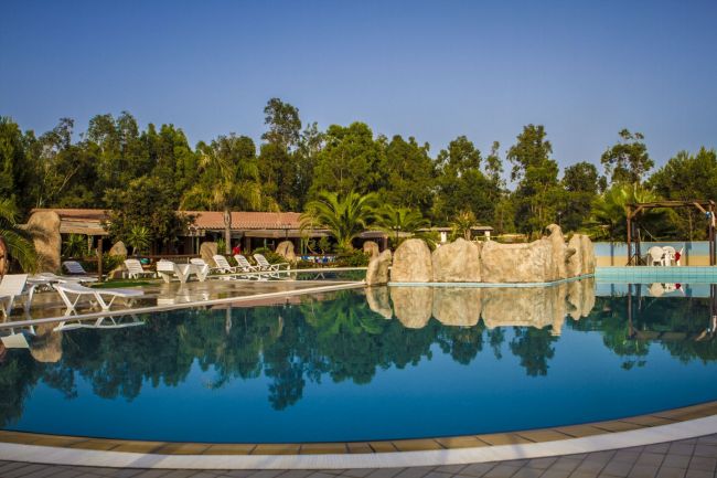 Piscina all'aperto con rocce decorative, circondata da lettini e alberi.