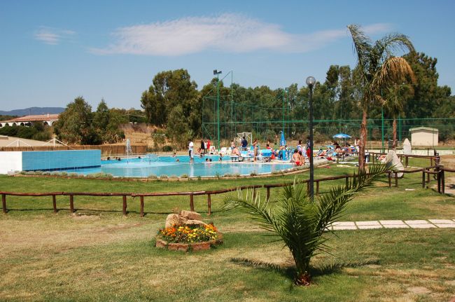 Una piscina all'aperto circondata da erba e alberi, con persone che si rilassano sui lettini.