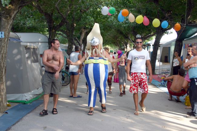 Persone in un campeggio, una indossa un colorato vestito da clown, palloncini appesi tra gli alberi.