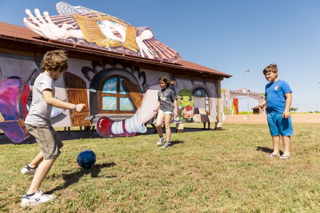 Tre bambini giocano a calcio davanti a murale colorato.