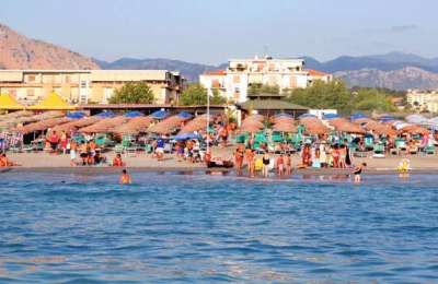 Una spiaggia affollata, con ombrelloni e lettini, e persone che nuotano nel mare.
