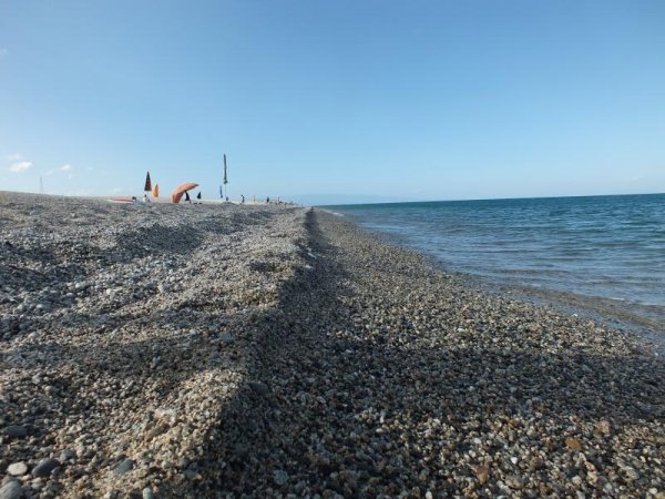Spiaggia di ciottoli con mare e alcune sdraio distanti.