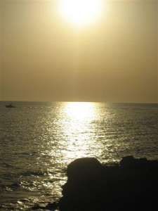 Sole al tramonto che riflette sull'acqua del mare, con silhouette di scogli.