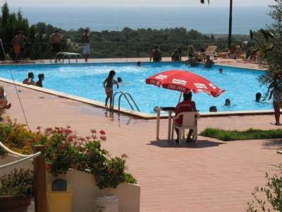 Piscina all'aperto con bagnanti, ombrellone rosso e vista mare.