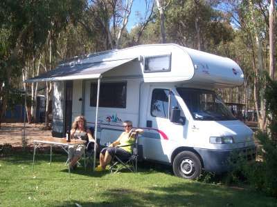 Due persone sedute davanti a un camper con tende aperte in un'area verde con alberi.