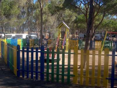 Parco giochi recintato con altalene e scivolo, circondato da alberi.