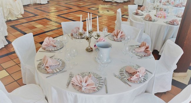 Tavolo rotondo apparecchiato con tovaglia bianca e tovaglioli rosa.