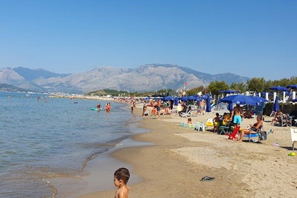 Spiaggia affollata con ombrelloni blu e montagne.