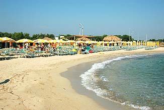 Spiaggia sabbiosa con mare calmo, lettini e ombrelloni gialli, capanne in lontananza.