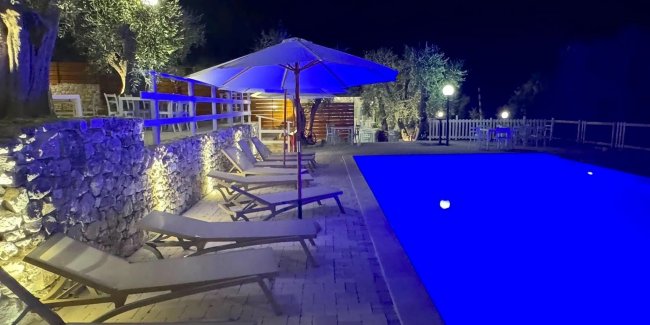 Piscina illuminata di notte con lettini e ombrellone.