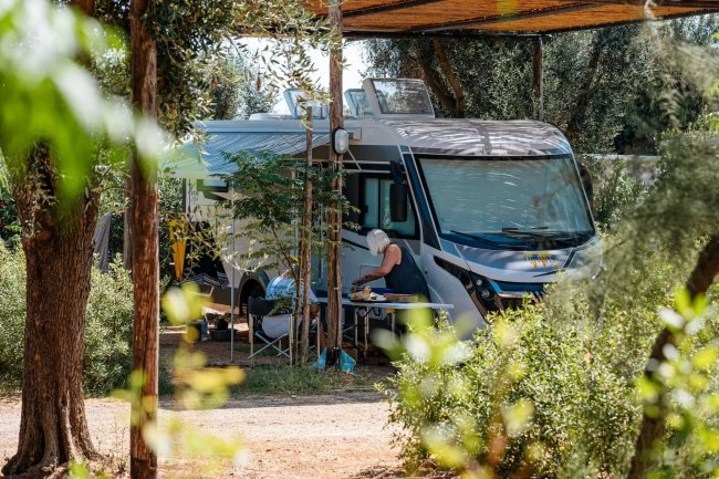 Camper parcheggiato tra gli alberi, persona seduta al tavolo.