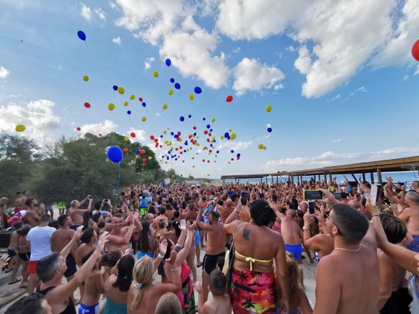 Una folla al mare lancia palloncini colorati in cielo. Giornata soleggiata con nuvole.