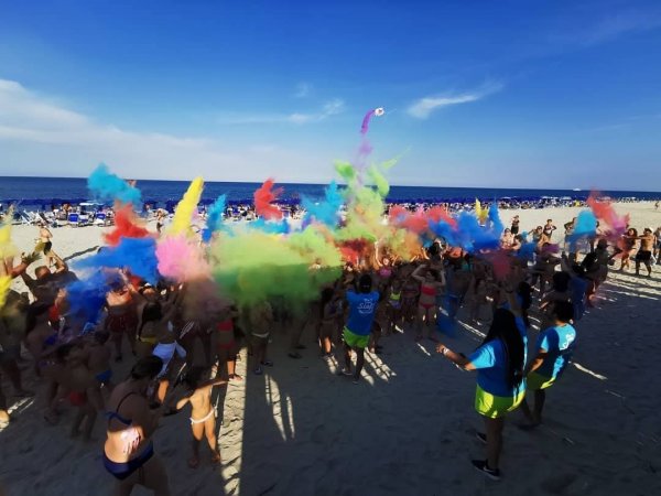 Spiaggia affollata con polveri colorate lanciate in aria durante un evento.