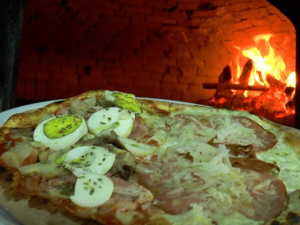 Pizza guarnita con uova e prosciutto cotto vicino a un forno a legna acceso.