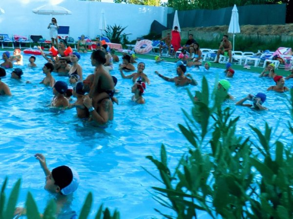Persone in una piscina all'aperto, alcuni bambini sulle spalle degli adulti.