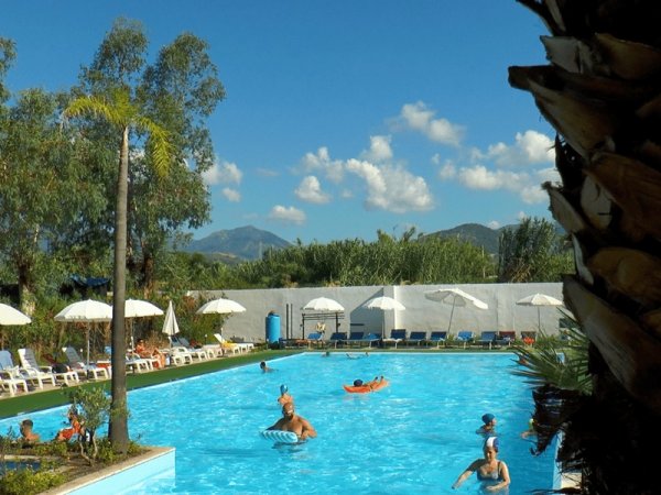Persone in piscina sotto il cielo azzurro con ombrelloni bianchi.