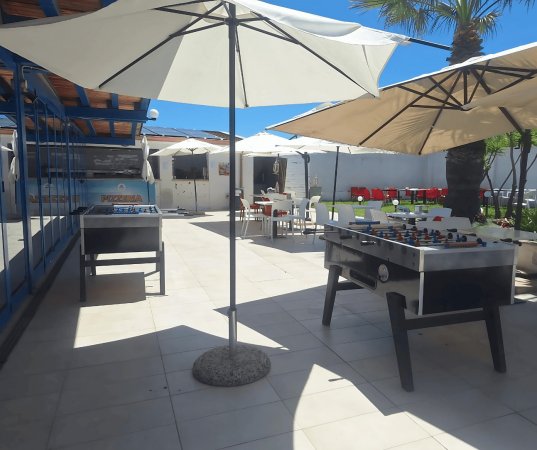 Tavoli da calcio balilla sotto ombrelloni in una terrazza soleggiata con sedie e tavoli all'aperto.