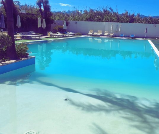 Grande piscina con acqua turchese e lettini intorno, ombrelloni chiusi e alberi sullo sfondo.