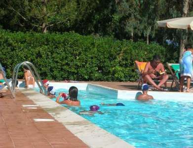 Piscina con bambini che nuotano e adulti seduti sotto ombrelloni.
