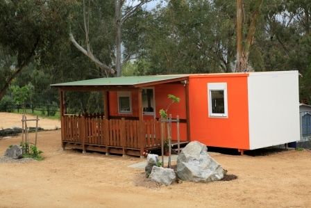 Una piccola casa prefabbricata arancione con veranda di legno e tetto verde in un'area alberata.
