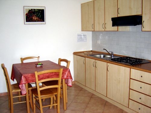 Cucina con mobili in legno chiaro, tavolo rettangolare coperto da tovaglia rossa, quattro sedie.