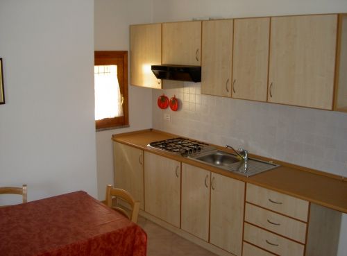 Cucina con armadi chiari, piano cottura, lavello e piccola finestra. Tavolo vicino.