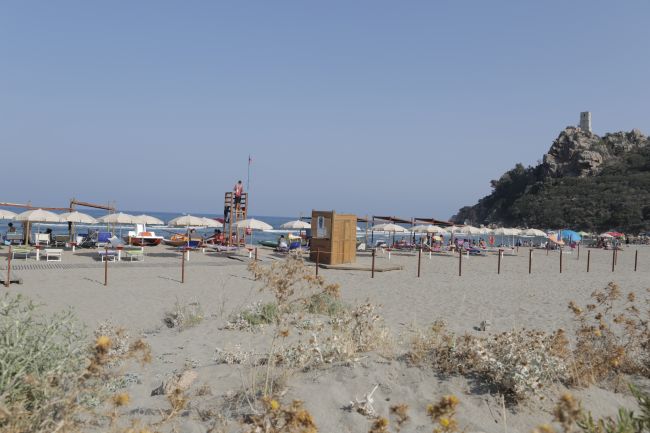 Spiaggia sabbiosa con ombrelloni e roccia collinare sullo sfondo.