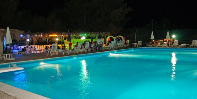 Piscina illuminata di sera, con sedie a sdraio e ombrelloni sul bordo.