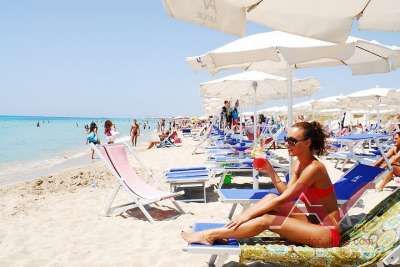 Spiaggia affollata con ombrelloni bianchi e sdraio, donna in costume sorseggia una bevanda rossa.