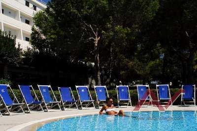 Una piscina con lettini blu, una persona in acqua e alberi sullo sfondo.