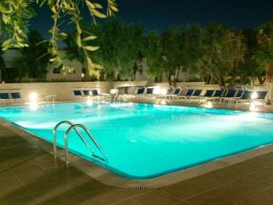Piscina illuminata di notte, con lettini intorno e alberi sullo sfondo.