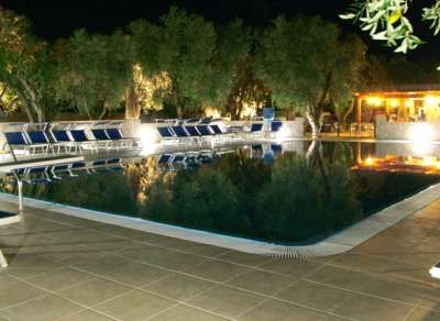 Piscina riflettente di notte, circondata da lettini, con alberi e bar illuminato.