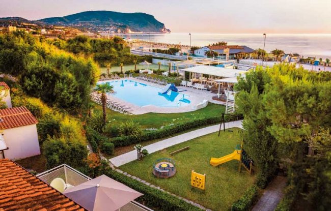 Vista su piscina e parco giochi, con mare e colline sullo sfondo.