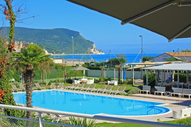 Piscina con sedie a sdraio, ombrelloni e vista sul mare e montagne.